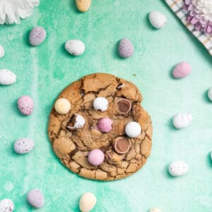 Overhead picture of a baked mini egg cookie surrounded by mini eggs and flower heads on a green/blue background.