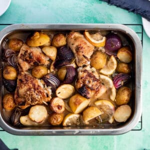 A tray of roasted chicken legs with potatoes and onions, freshly cooked and served on a light blue table.