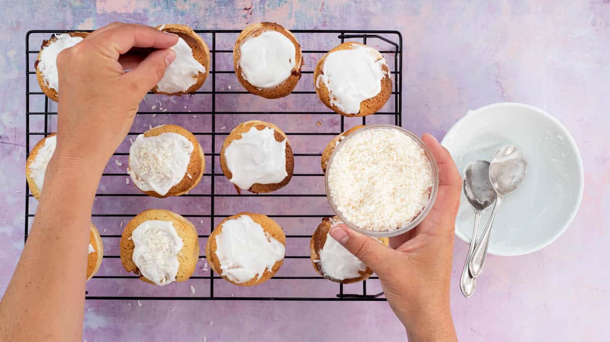 A photo showing hands sprinkling desiccated coconut over the baked and iced London Cheesecakes while they sit upon a cooling rack.