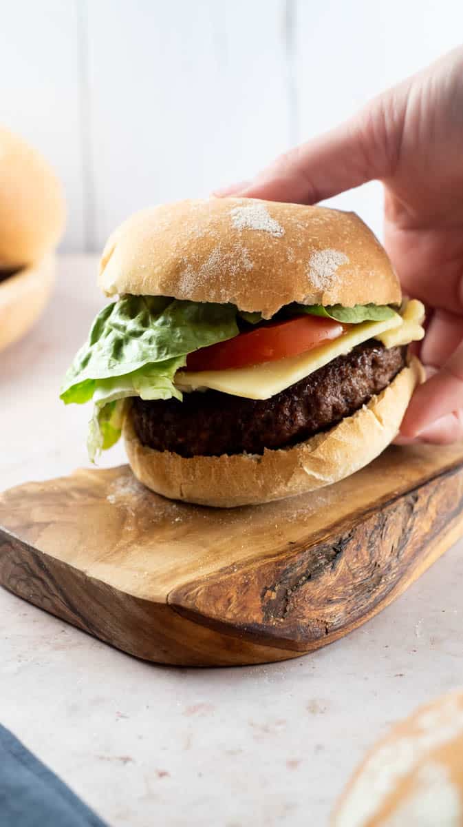 A hand is lifting a filled burger bun from a wooden board.
