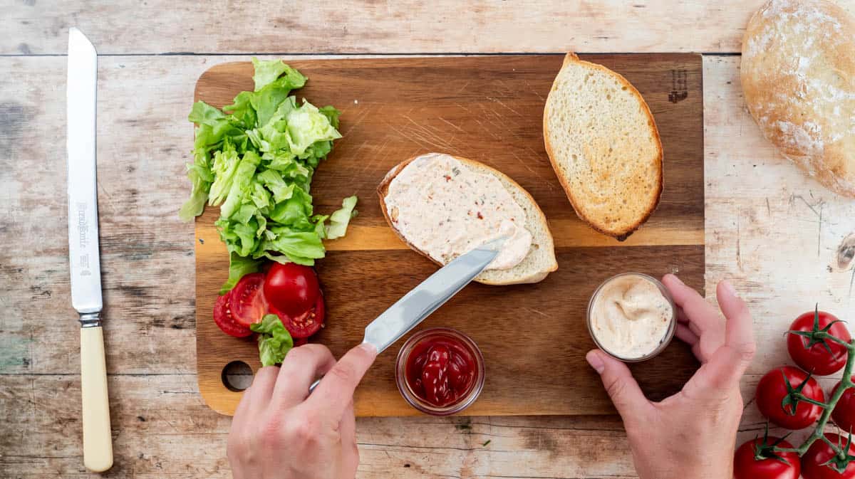 A hand spreading one side of a toasted Portuguese bun with Peri Peri mayonnaise.