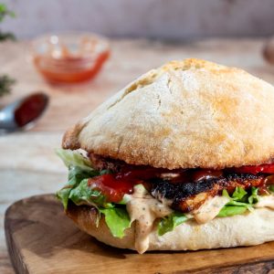 A Butterfly chicken burger sat on a wooden board with Perinaise sauce spilling out of the side.