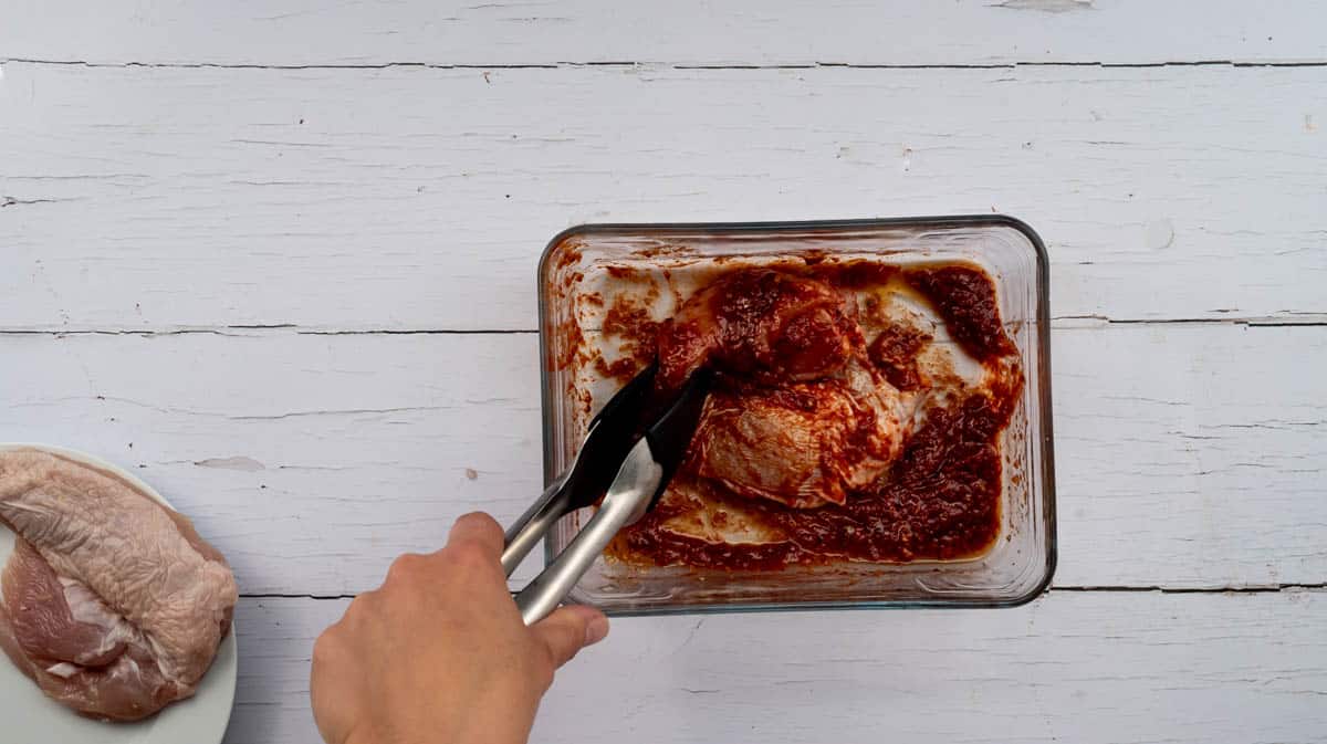 A hand holding tongs to move the chicken breast and coat it in the marinade.