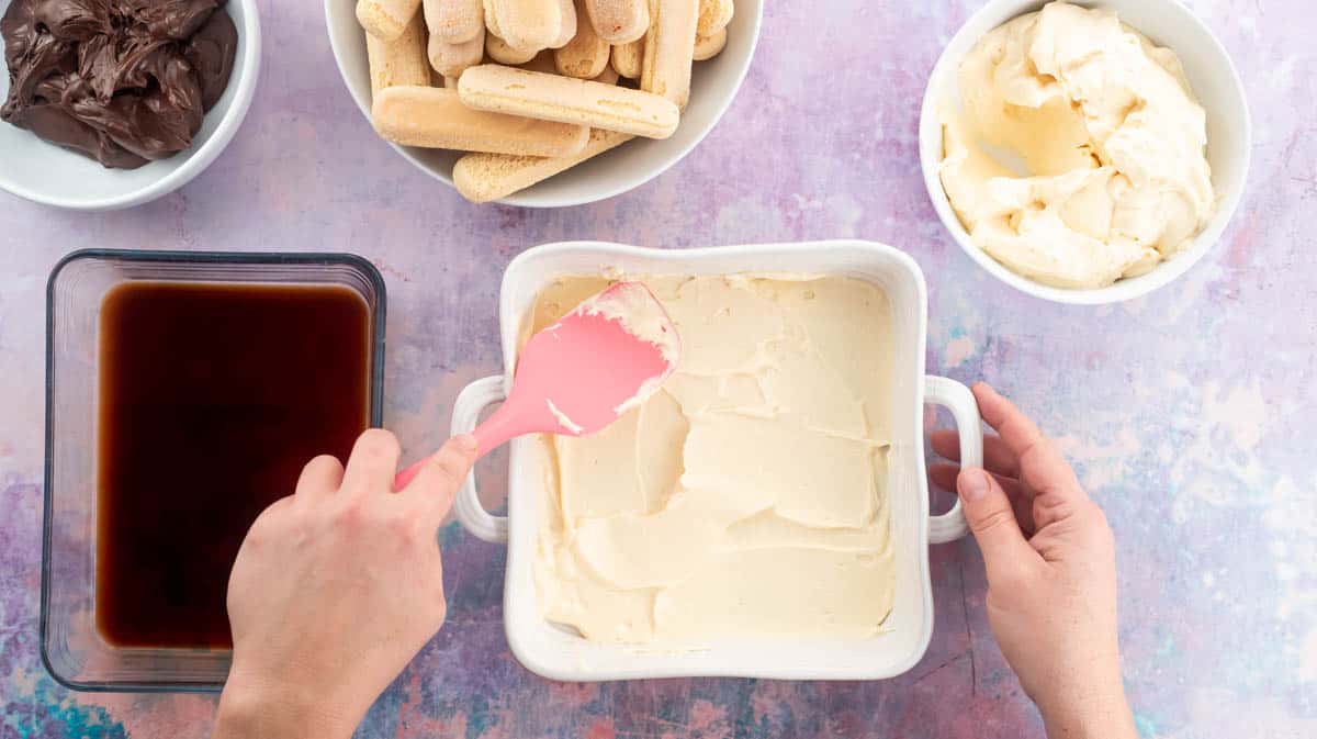 A hand is using a pink spatula to spread a layer of mascarpone cream over the bottom of a white serving dish.