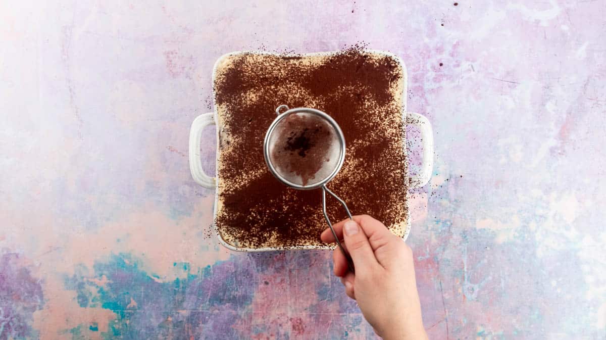 A hand is using a small sieve to dust the surface of the Nutella Tiramisu with cocoa powder.
