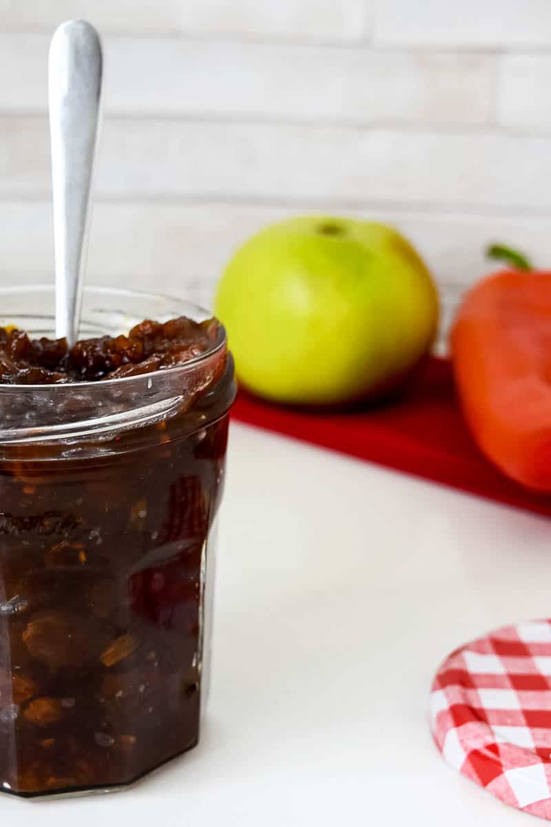 A close up of a jar filled with Autumn Chutney with a spoon in it. In the background there are ingredients.