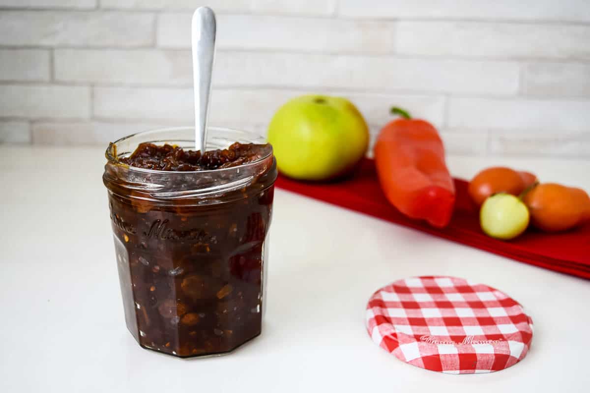 A jar of Chutney with a spoon inside. There is an apple, pepper, and tomatoes in the background.