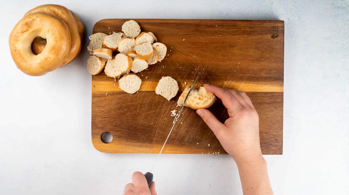 A hand is using a serrated knife to slice bagels on a chopping board.