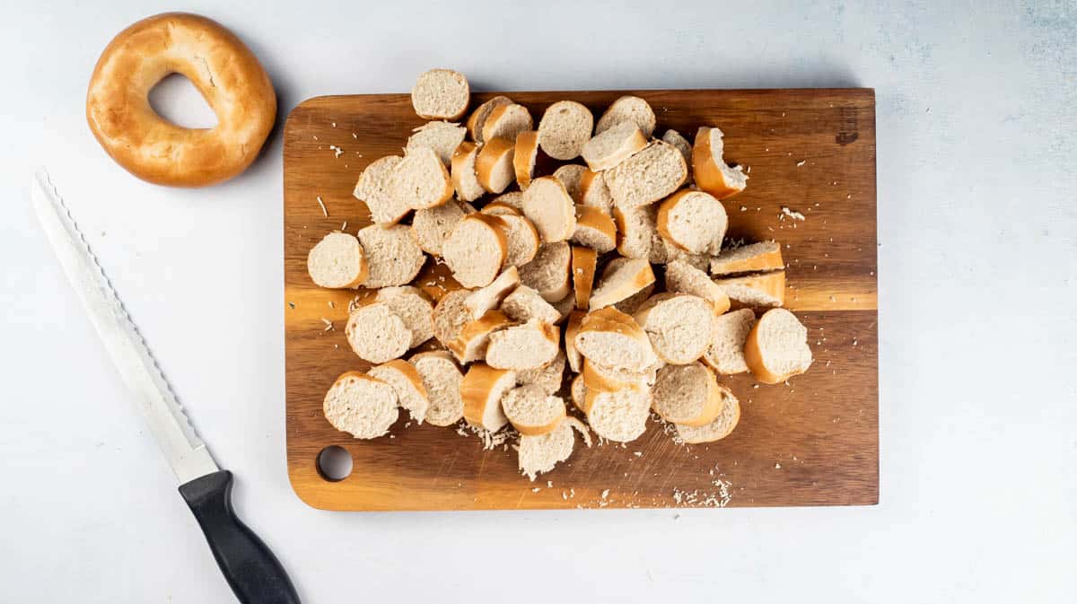 A pile of sliced bagels on a wooden board with a whole bagel and a serrated knife next to it.