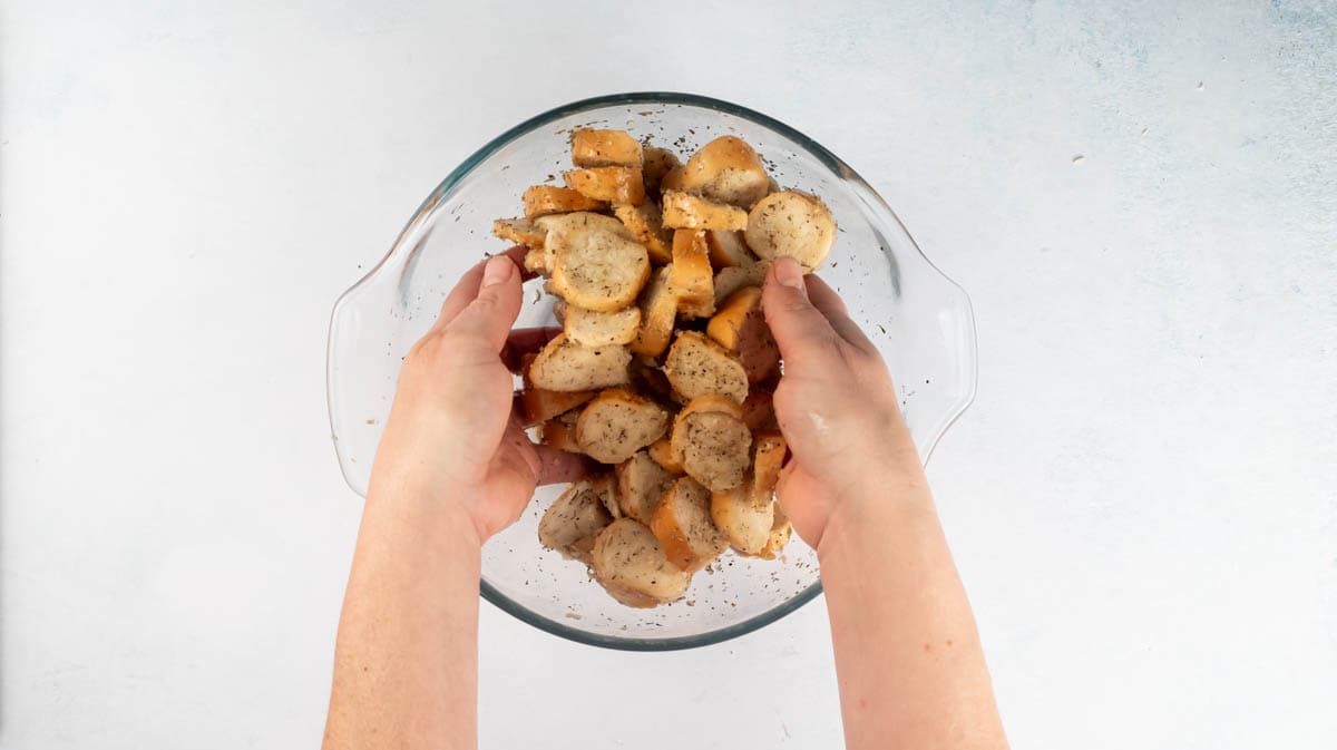 Two hands are tossing bagel slices in seasoned oil inside a glass bowl.