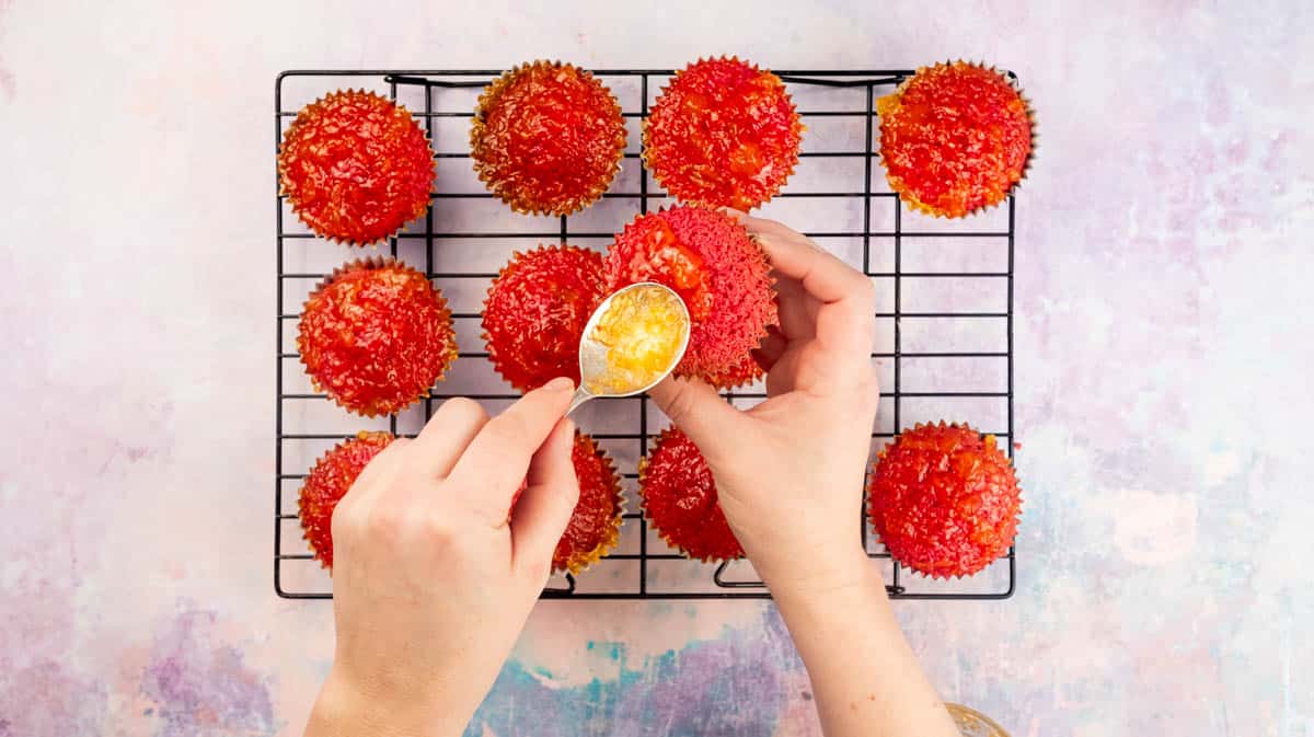 A hand is using a spoon to cover the top of cupcakes with apricot jam.