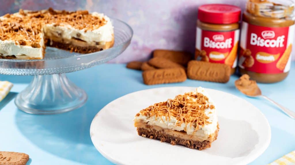 A front view of a slice of Banoffee Pie on a white plate. In the background there is the rest of the pie on a cake stand, some Biscoff biscuits and two jars of Biscoff spread.