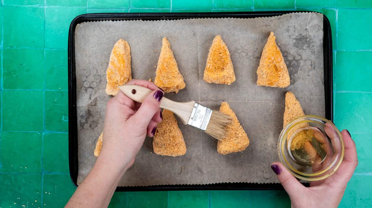A hand is using a pastry brush to brush camembert wedges with oil on a lined baking tray.