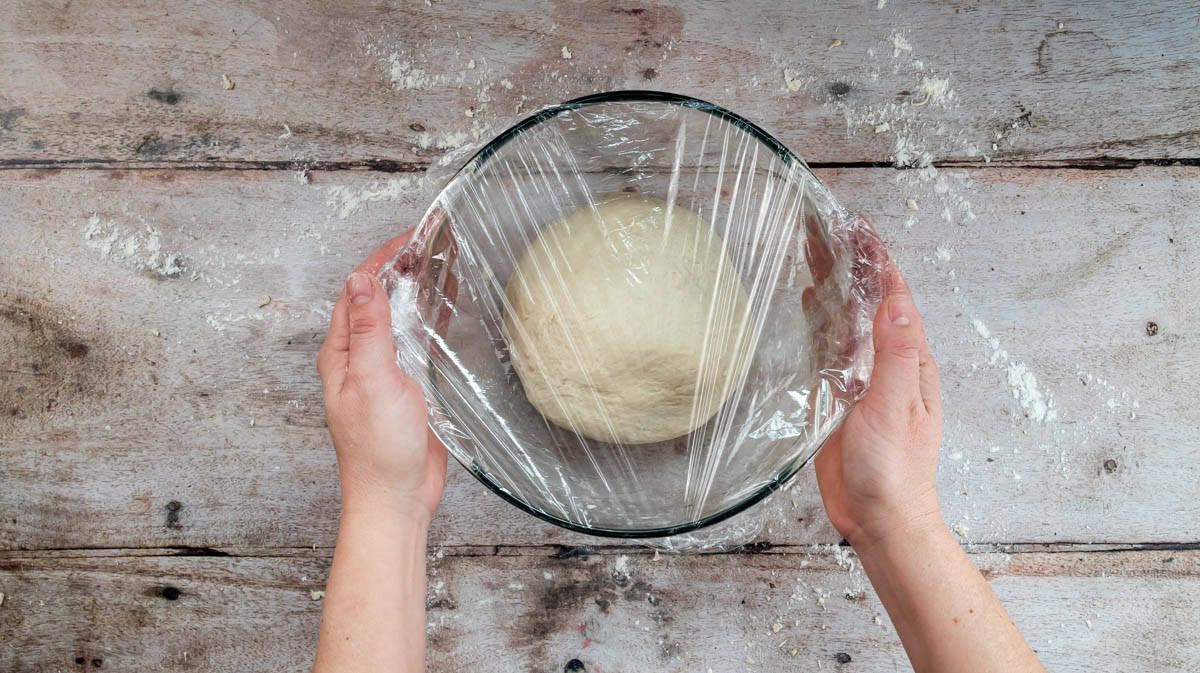 Two hands are holding a glass bowl with a bread dough ball inside, that has been covered with cling film.