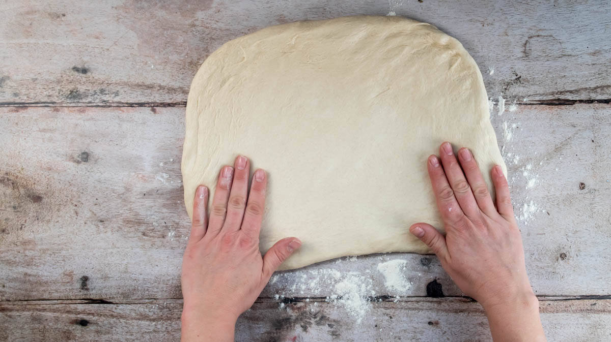 Two hands are flattening bread dough into a rectangle shape on a flowered surface.