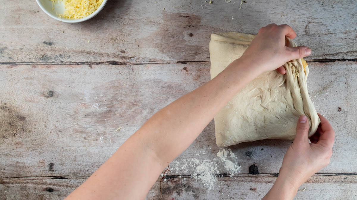 Two hands are completing the final fold of the bread dough and sealing the edges.