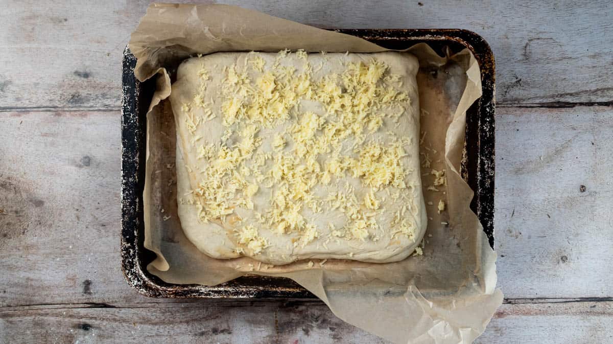 A rectangular bread dough placed inside a lined baking tin and sprinkled with cheddar cheese.