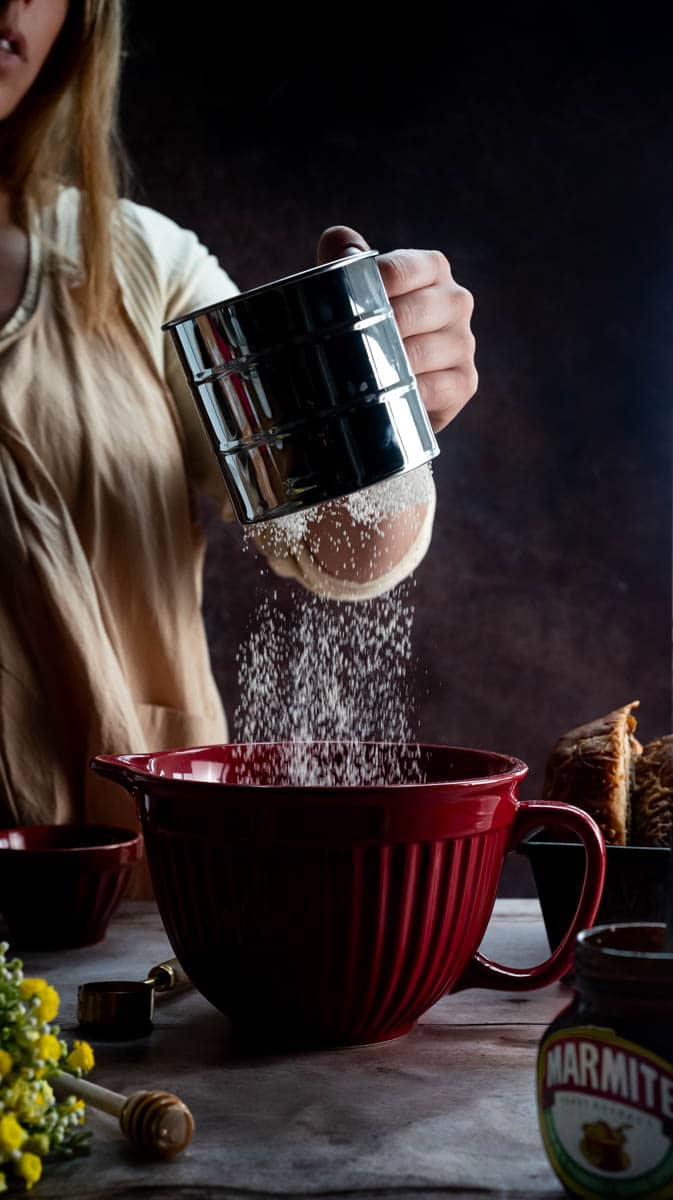 A person is sifting flour into a red jug shaped bowl. There are decorative ingredients and items surrounding the bowl.