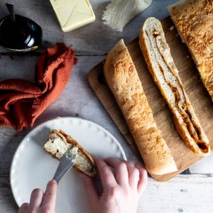 Overhead view of a loaf of cheese and marmite bread with a hand spreading a slice with butter.