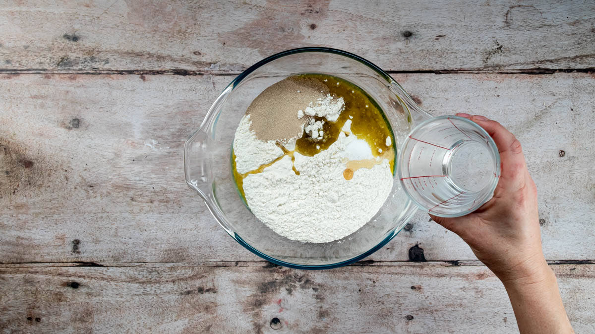 A hand is tipping water into a glass bowl that contain flour, yeast, salt, honey and oil.
