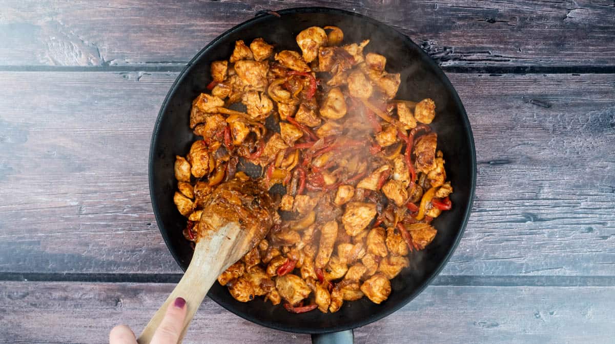 A hand is using a wooden spoon to stir the chicken mixture as it fries.
