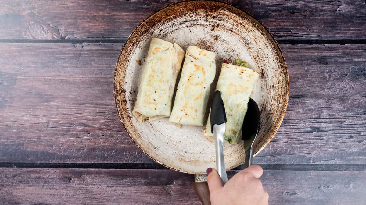 A hand is using tongs to turn the folded wraps over in a frying pan as they toast on both sides.