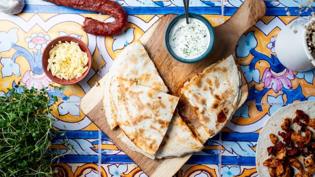 A chorizo quesadilla sliced into wedges and stacked on a wooden board with garlic Mayo next to them. Surrounded by decorative items and ingredients.