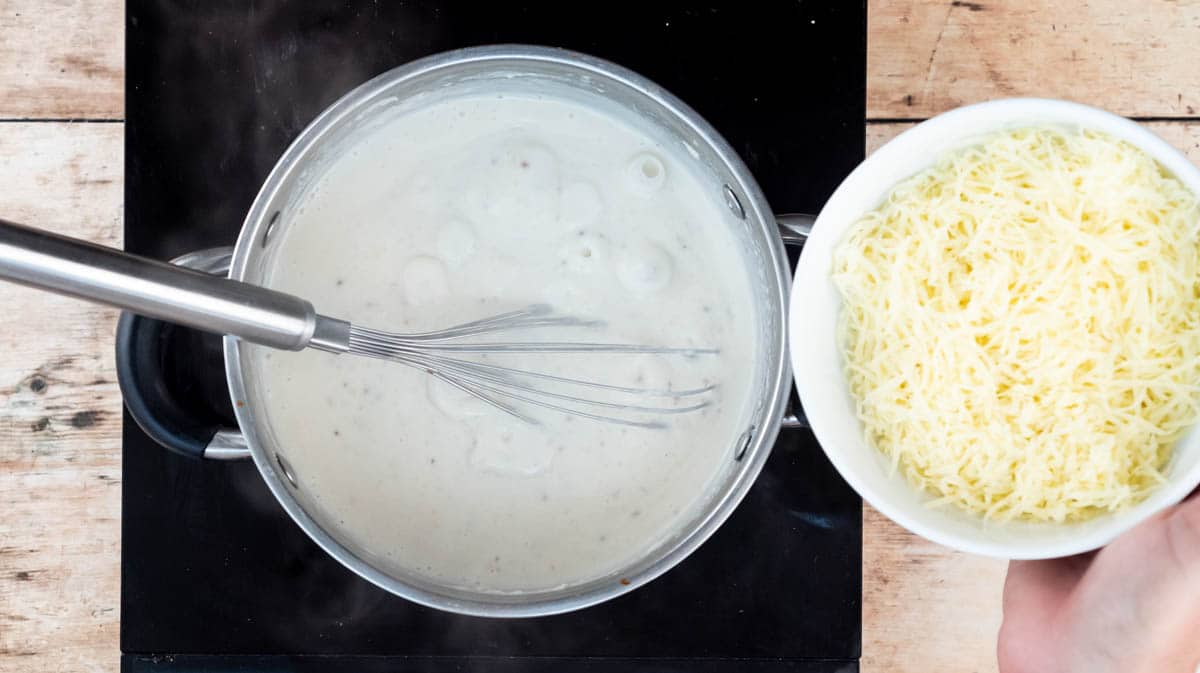 A hand is adding Gruyere cheese into the white sauce which is bubbling in a saucepan.
