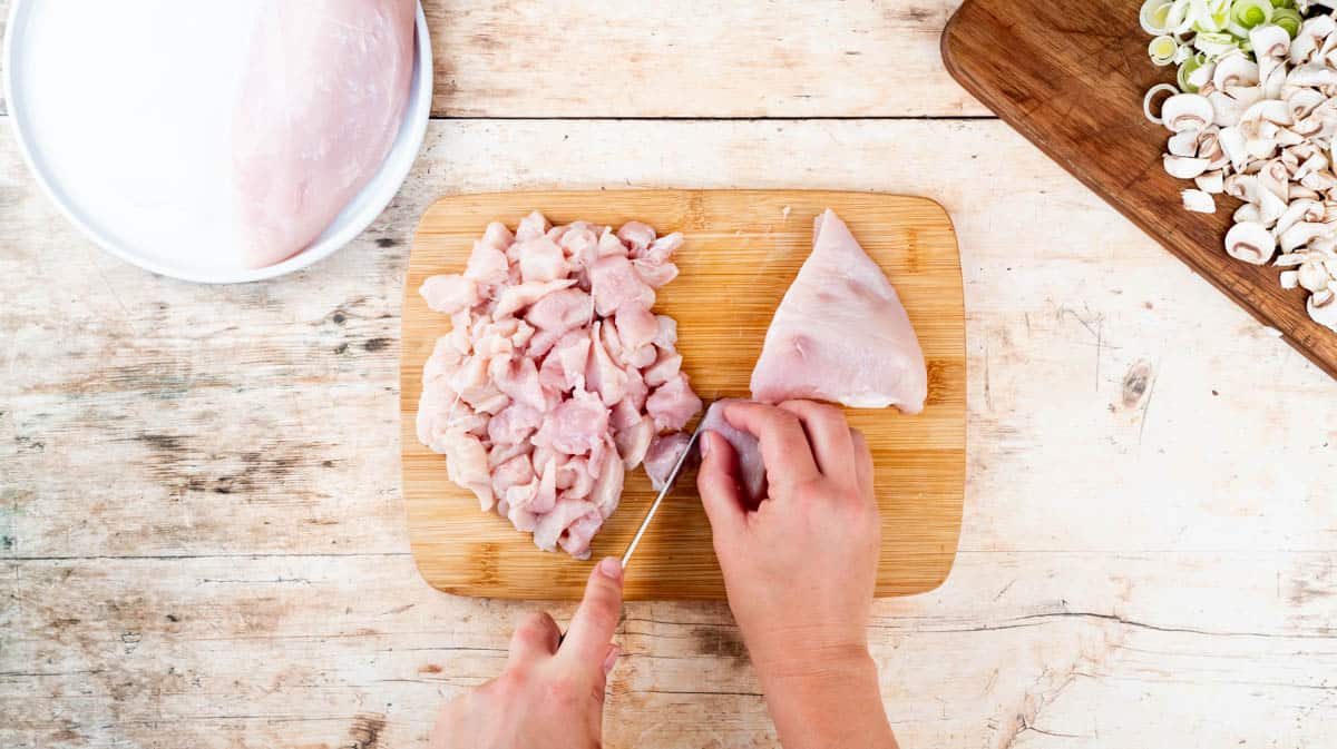 A hand is using a sharp knife to dice chicken into small pieces.