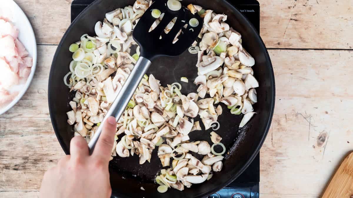 A hand is using a spatula to mix sliced mushrooms and leeks in a frying pan.