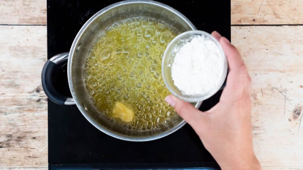 A hand is tipping flour into a saucepan containing melted butter.