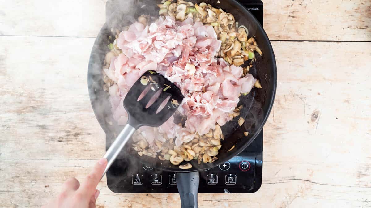 A hand is using a spatula mix diced chicken and streaky bacon pieces into the cooked mushrooms and leeks in a frying pan.