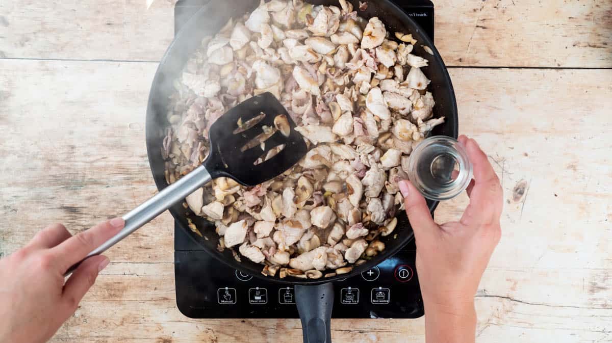 A hand is adding white wine to the ingredients in a frying pan to deglaze it.