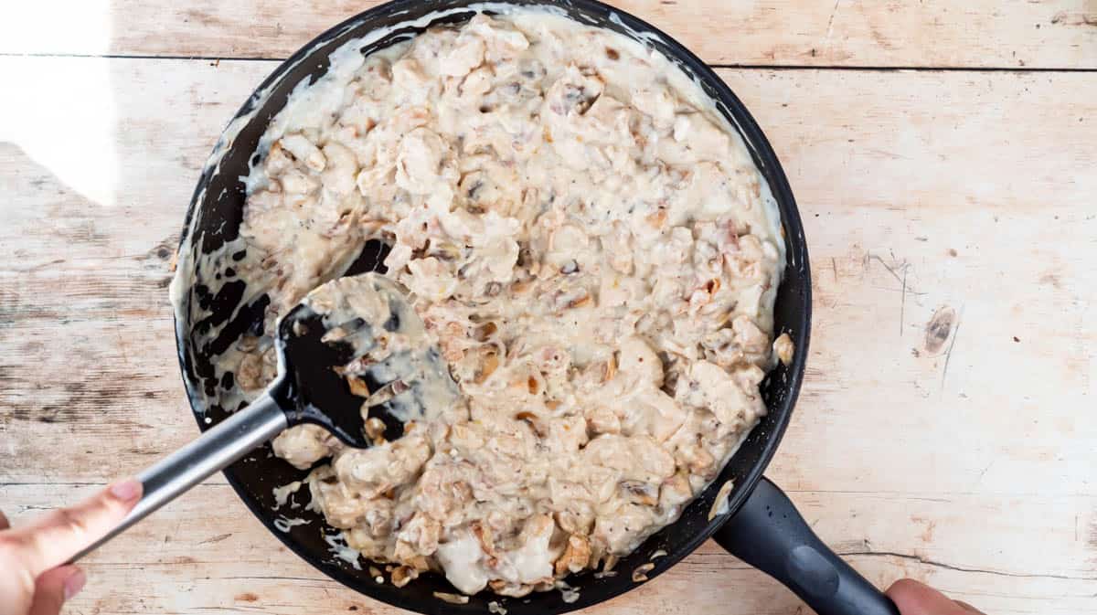 A hand is using a spatula to mix the white sauce in with the chicken and mushroom mixture in a frying pan.