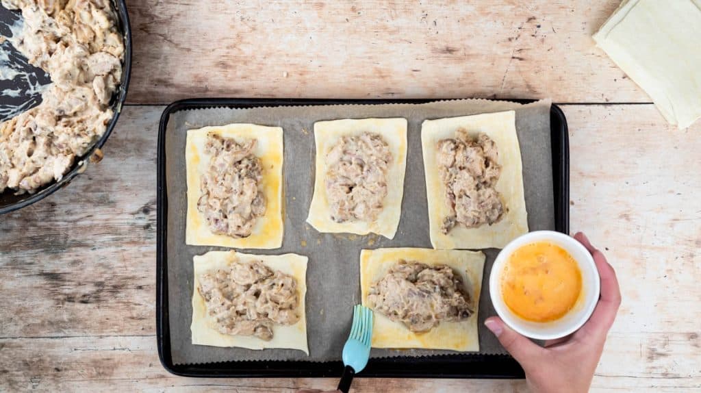 A hand is using a pastry brush to brush beaten egg around the sides of the filling on a rectangle of puff pastry.