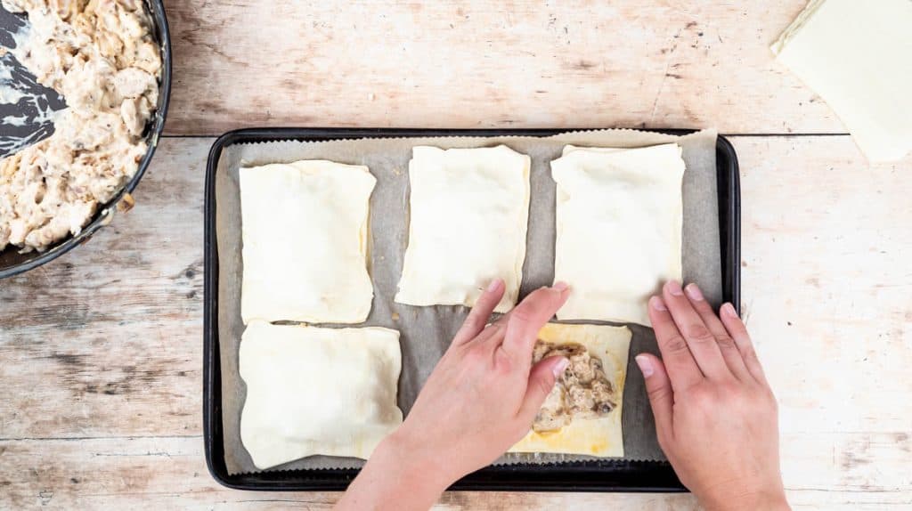 Two hands are placing a rectangle of puff pastry on top of another piece that has the filling in the middle.