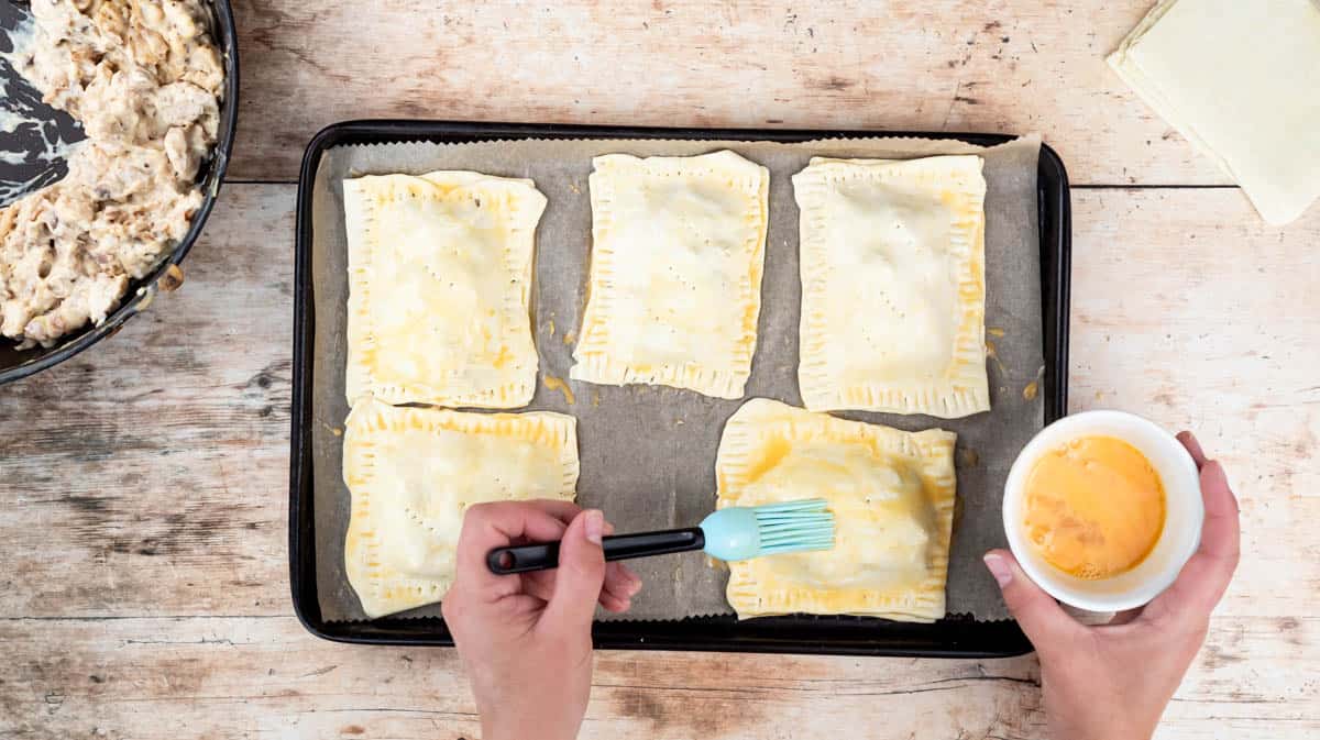 A hand is using a pastry brush to brush beaten egg on the tops of the chicken and mushroom pasties.