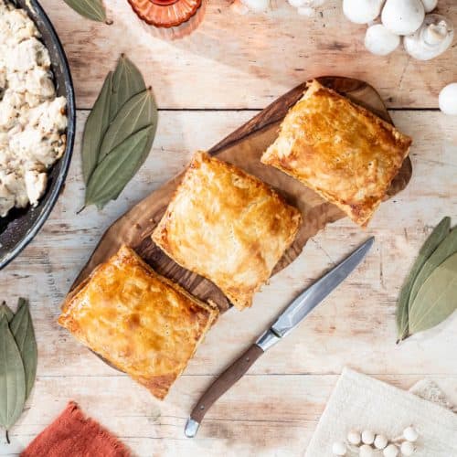 Three chicken and mushroom pasties on a wooden board surrounded by ingredients and decorative items.