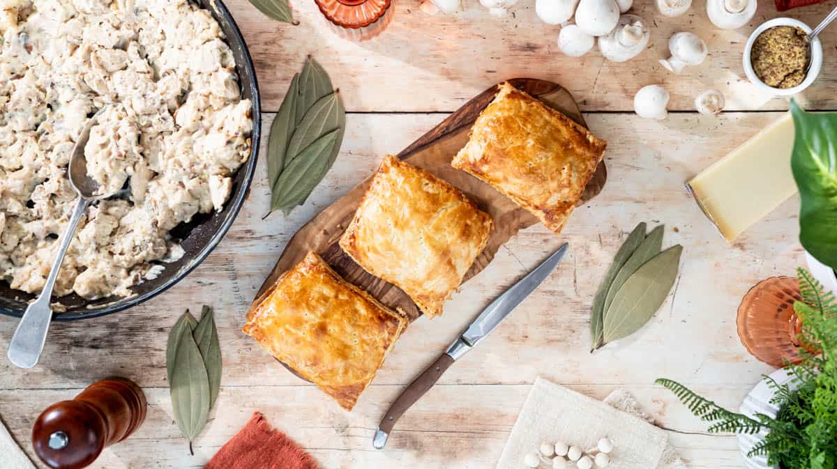 Three chicken and mushroom pasties on a wooden board surrounded by ingredients and decorative items.
