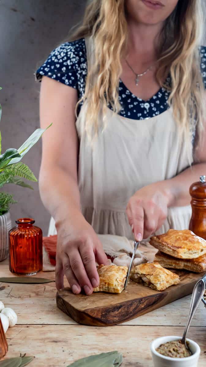 A person is slicing a chicken and mushroom pasty in half on a wooden chopping board.