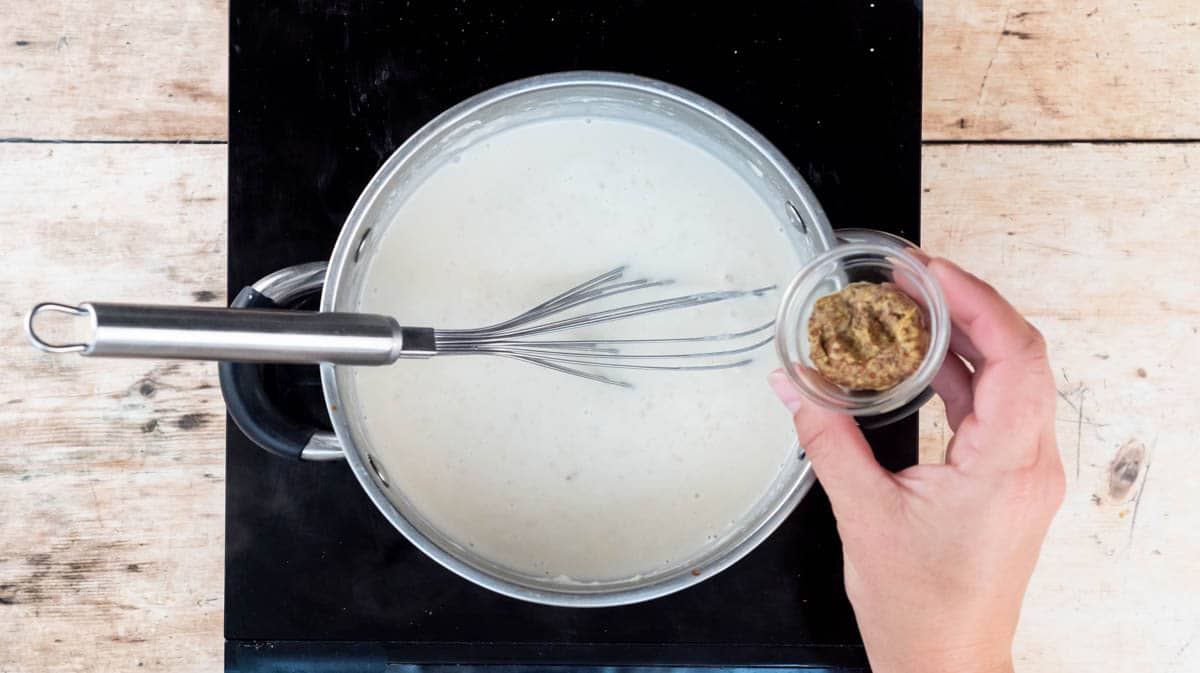 A hand is adding wholegrain mustard to the white sauce in a saucepan.