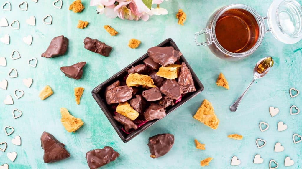 An overhead view of Cinder Toffee piled into a rectangle tin surrounded by more pieces.