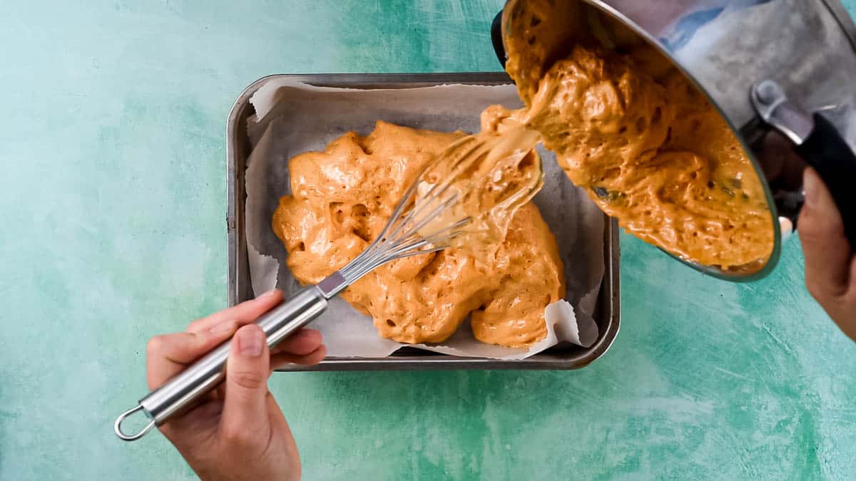 A hand is using a whisk to transfer the mixture to a lined baking tin.
