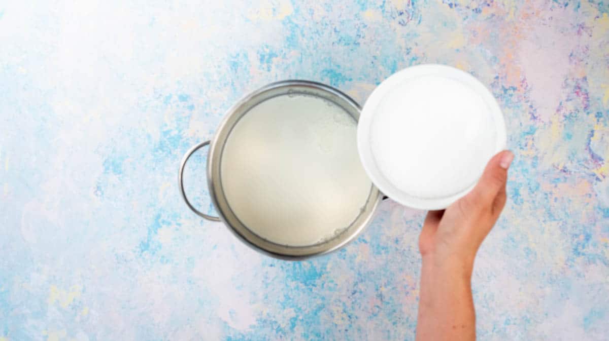 A hand is pouring caster sugar into a saucepan that is filled with milk.