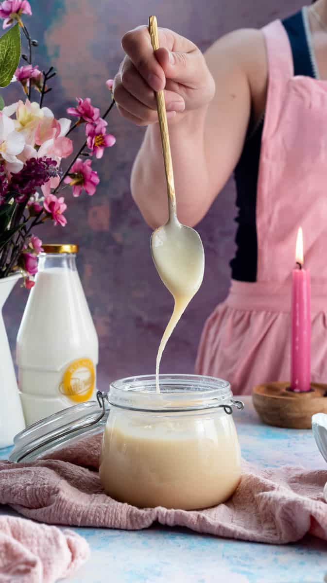 A person is lifting a gold spoon from the jar of condensed milk as it drizzles from the spoon back into the jar.