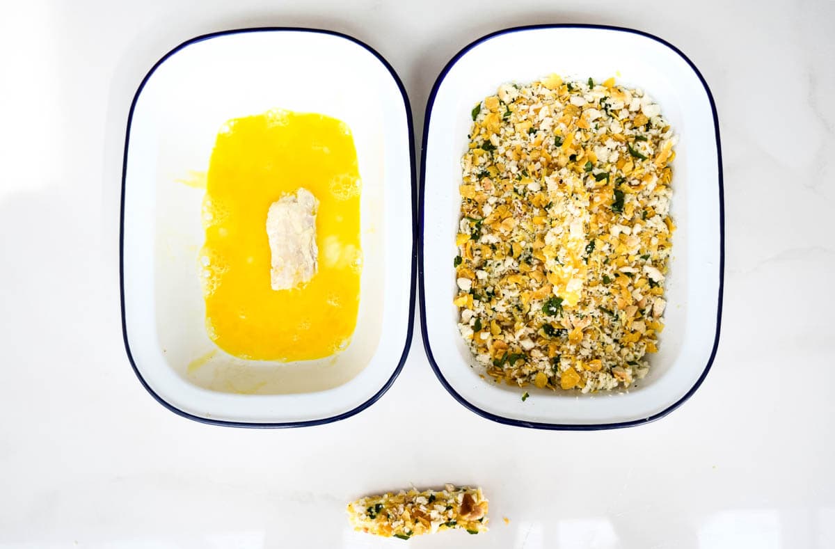 Two bowls, one with beaten egg and a piece of cod inside. The other with the cornflake coating and another piece of cod. Below, there is a coated fish finger.