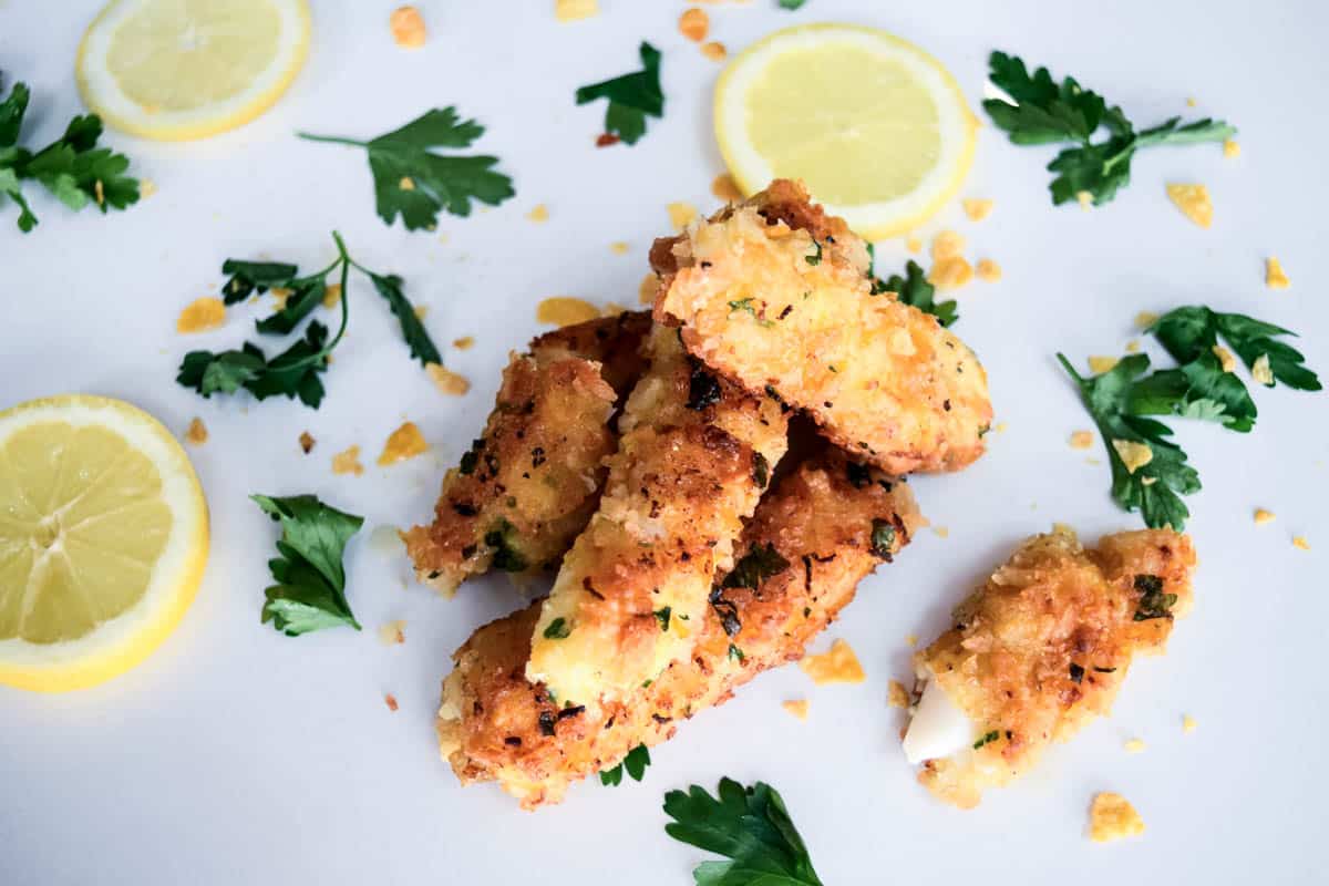An overhead view of a pile of Cornflake coated fish fingers with fresh parsley leaves and lemon slices surrounding them.