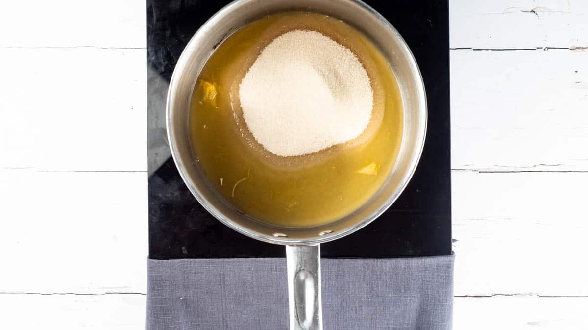An overhead view of a large saucepan on a stove with orange juice and caster sugar inside.