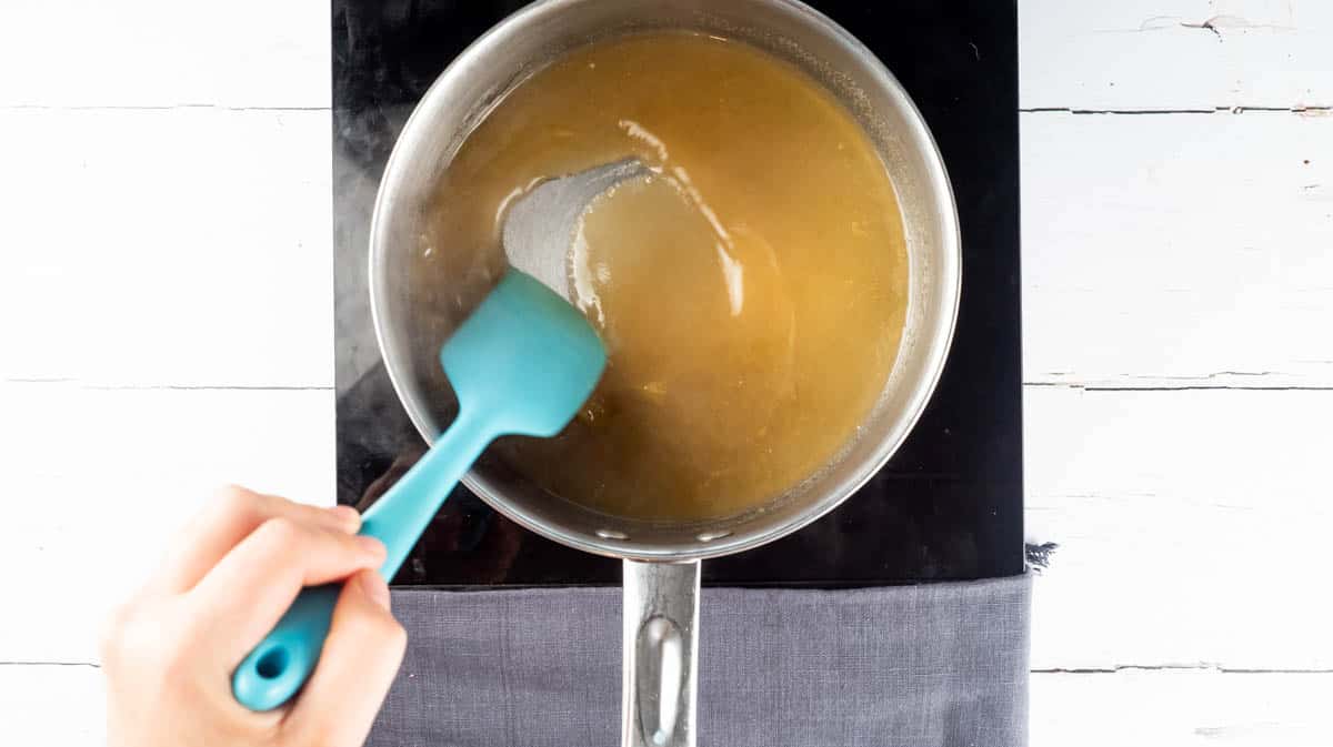 An overhead view of a hand stirring the sugar and orange juice together inside a saucepan.