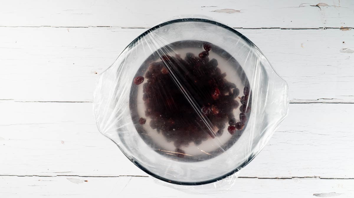 The cranberries soaking in a glass bowl that has be covered in clingfilm.